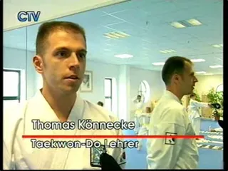 Screenshot Chiemgau TV vom 28. Mai 2003: Thomas Könnecke im Dobok im Interview, Namensbanner „Thomas Könnecke, Taekwon-Do Lehrer", im Hintergrund trainierende Schüler in Doboks
