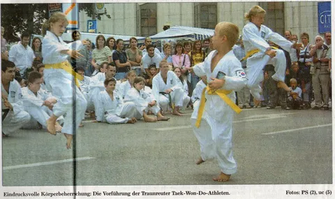 Foto Traunreuter Anzeiger vom 14. Juli 2003: Kinder mit gelben Gürteln bei der Taekwon-Do-Vorführung auf dem Traunreuter Stadtplatz, ein Mädchen springt, sitzende Mitschüler und Publikum im Hintergrund