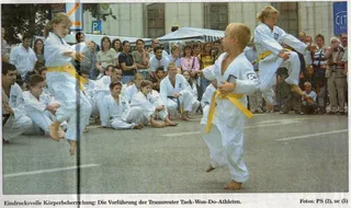 Foto Traunreuter Anzeiger vom 14. Juli 2003: Kinder mit gelben Gürteln bei der Taekwon-Do-Vorführung auf dem Traunreuter Stadtplatz, ein Mädchen springt, sitzende Mitschüler und Publikum im Hintergrund
