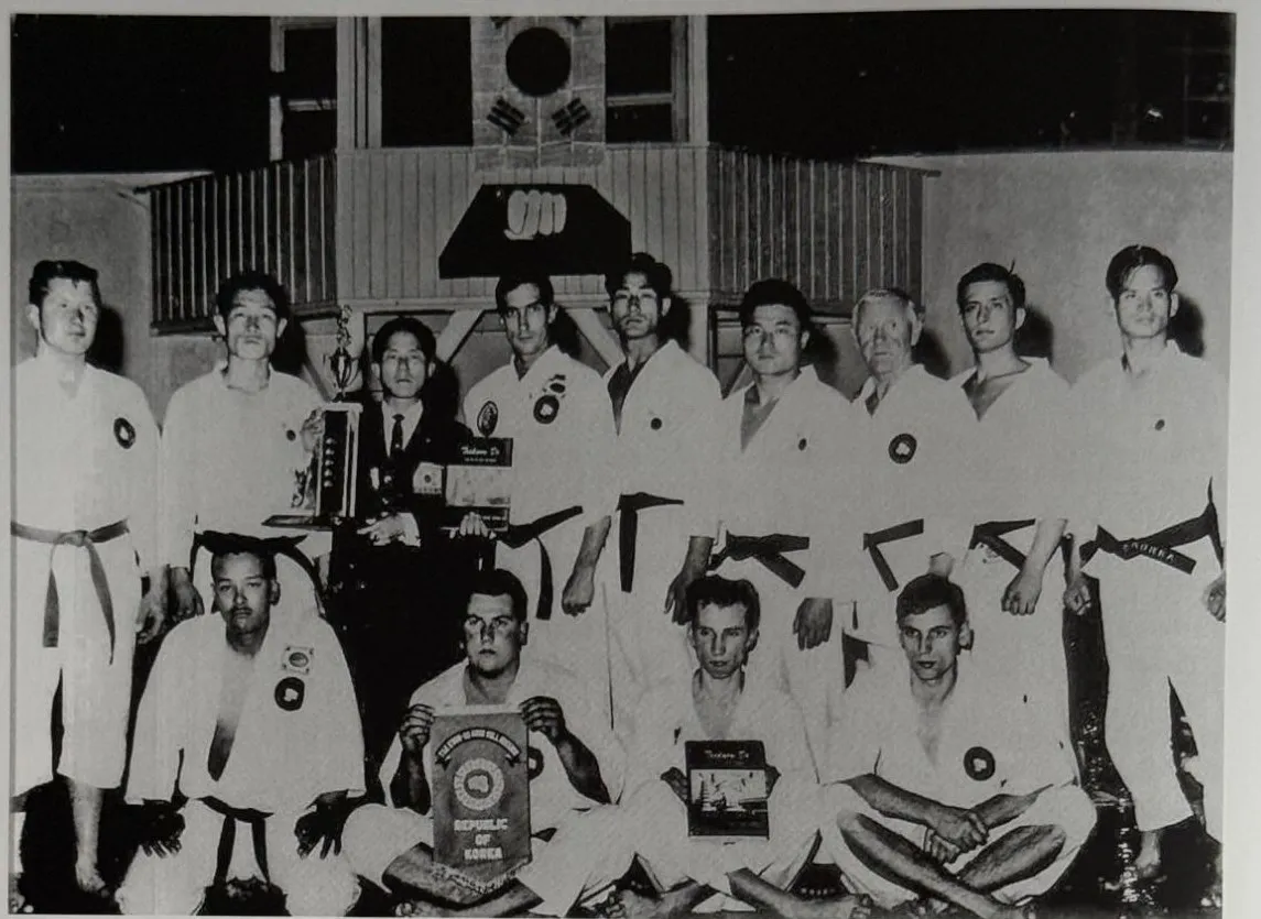 Gruppenfoto in einer Turnhalle unter der koreanischen Flagge: neun Männer in weißen Anzügen stehen, vier sitzen davor, in der Mitte Choi Hong-Hi im Anzug mit Pokal, Garmisch-Partenkirchen 1965