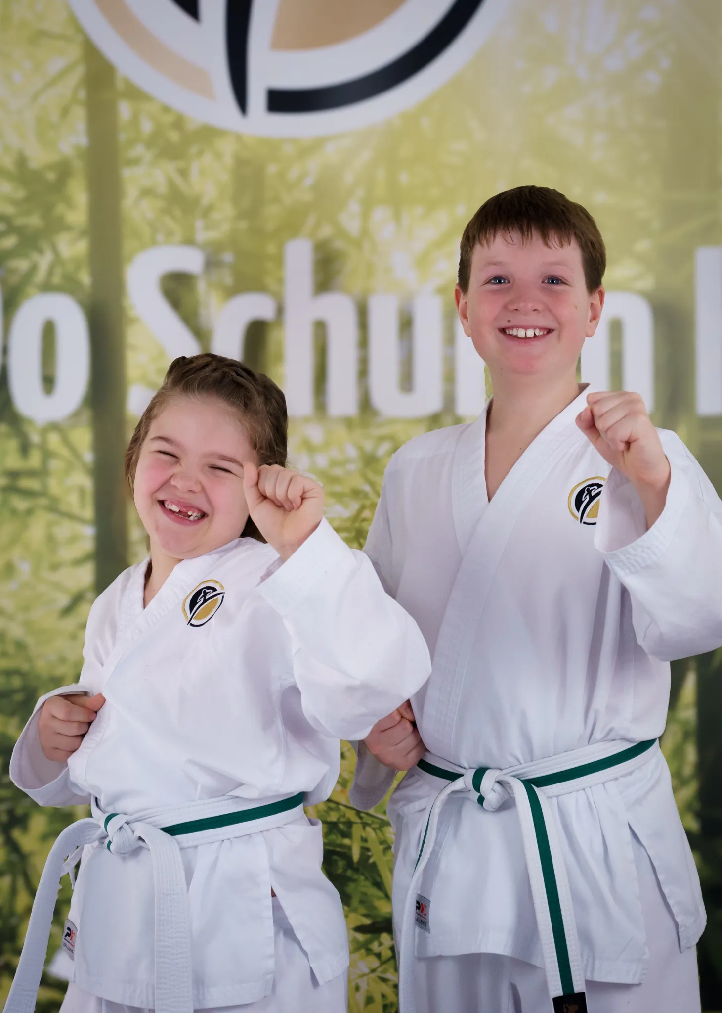 Zwei lächelnde Grundschulkinder im weißen Dobok mit erhobenen Fäusten posieren vor einem Banner der Taekwon-Do Schulen Könnecke