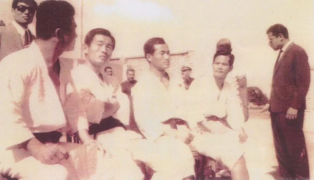 Teammitglieder im Dobok sitzen am Boden, Gastgeber in Anzügen und Uniformen stehen dahinter