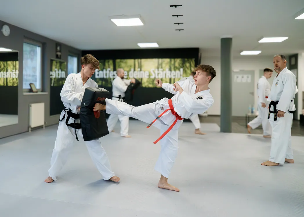 Zwei Jugendliche in weißen Doboks üben Pratzentraining, einer haltend, einer kickend, im Hintergrund beobachtet ein Trainer weitere Trainierende
