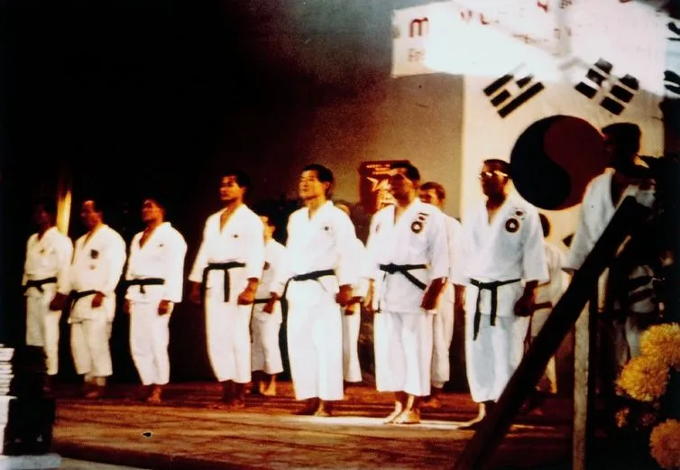 Farbfoto: Männer im Dobok stehen in einer Reihe auf einer Bühne vor koreanischer Flagge