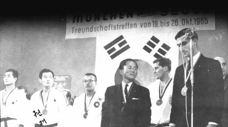 Männer im Dobok mit Medaillen und ein Redner am Mikrofon vor dem Banner „München grüßt Seoul“