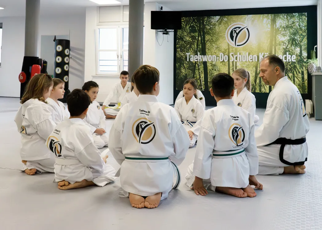 Kinder im Dobok sitzen aufmerksam im Sitzkreis, eine Trainerin leitet die Runde, im Hintergrund ist das Vereinsbanner der Taekwon-Do Schulen Könnecke zu sehen