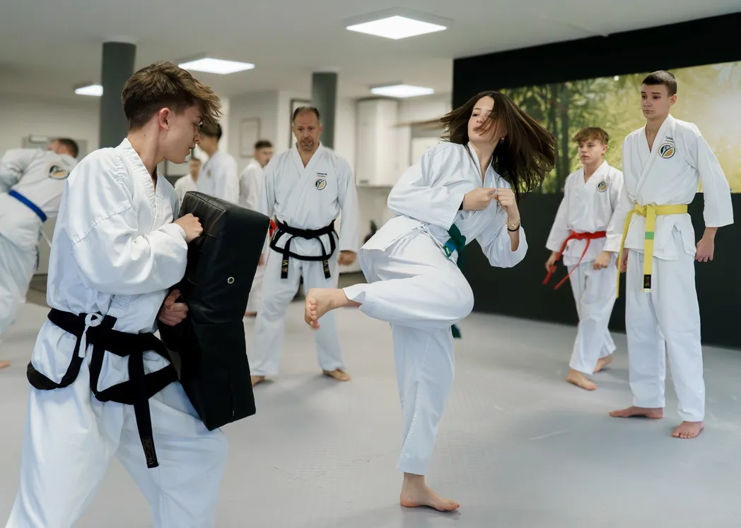 Jugendliche im weißen Dobok führt einen konzentrierten Kick gegen ein Pratzenpolster aus, das ein Trainingspartner hält, im Hintergrund beobachten weitere Jugendliche und ein Trainer